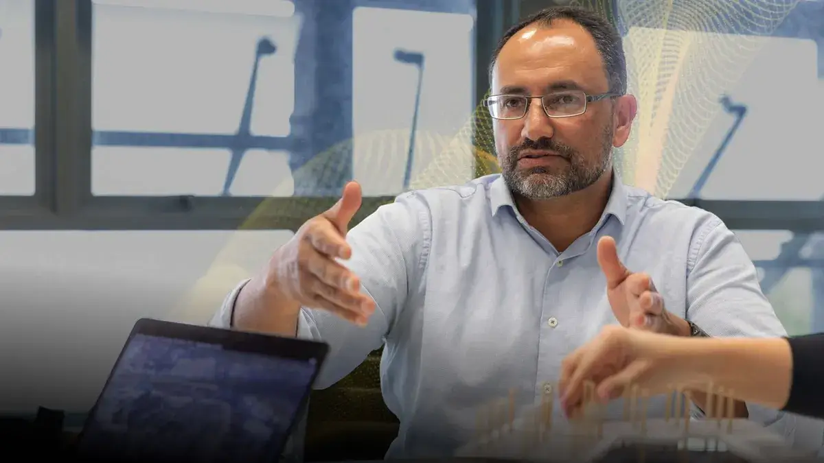 A man in glasses is gesturing while engaged in a conversation. He's seated at a table with a laptop, depicting a collaborative and focused atmosphere.
